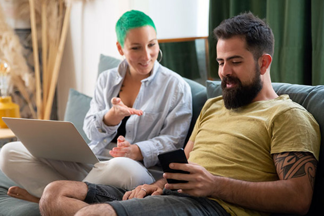 man using a phone sitting beside woman using a laptop on a couch at home