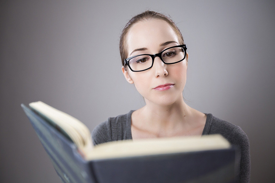 Woman with glasses reading a book in front of a gray backdrop