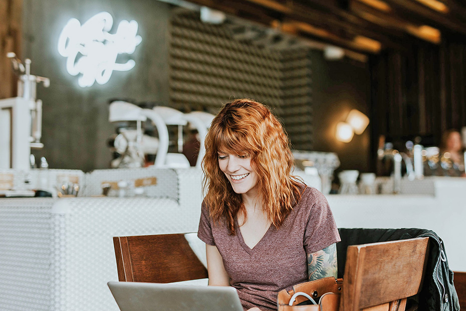 Red haired female in a coffee shop happily working on a laptop