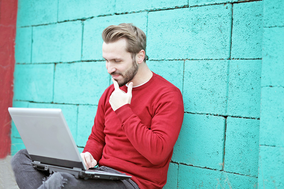 Happy adult male outside on laptop sitting against a teal colored wall