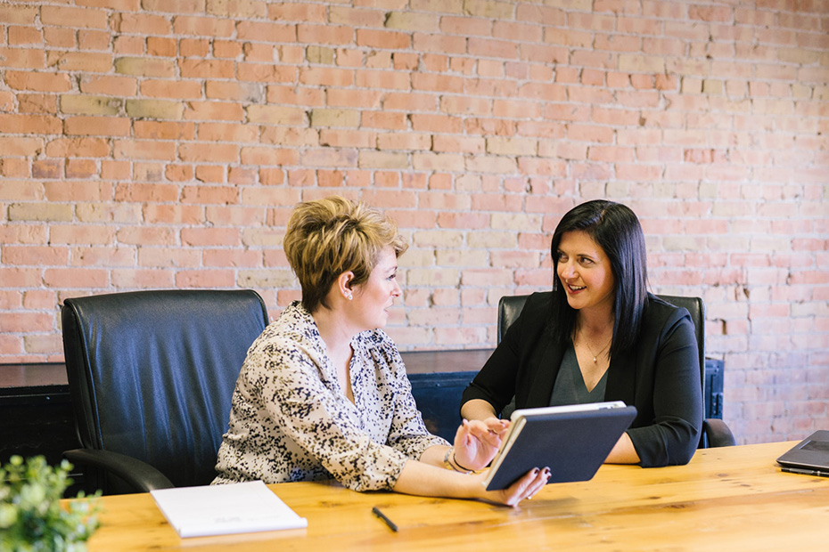 Two women in buiness casual clothes have a meeting in a meeting room, the woman on the left is holding a tablet