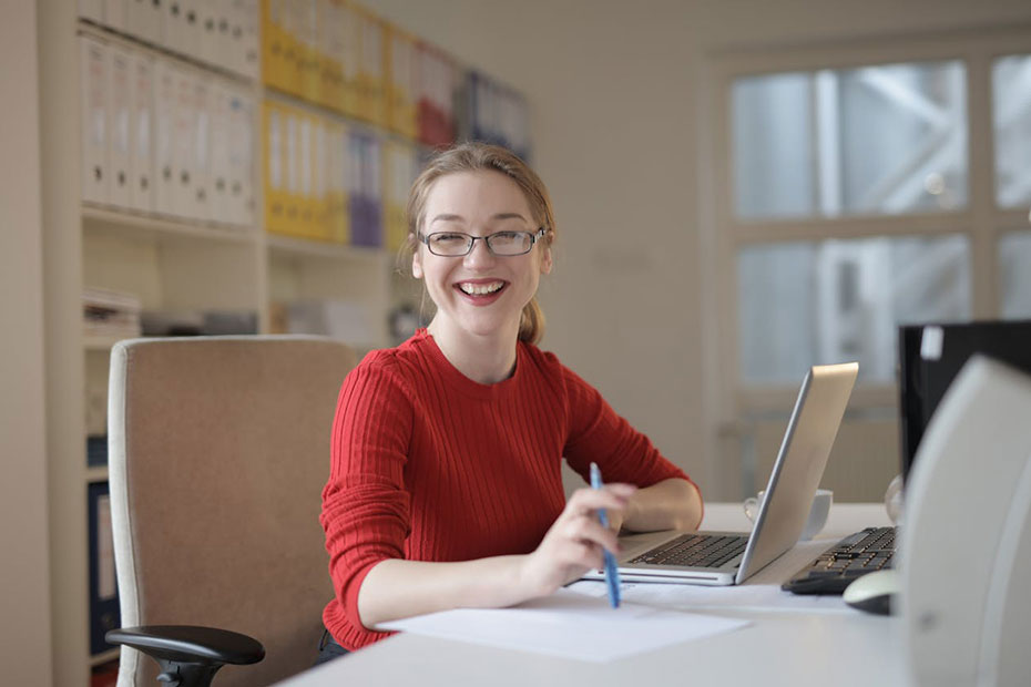 Smiling woman wearing glasses and a red long sleeve t-shirt working a ta a desk on a laptop