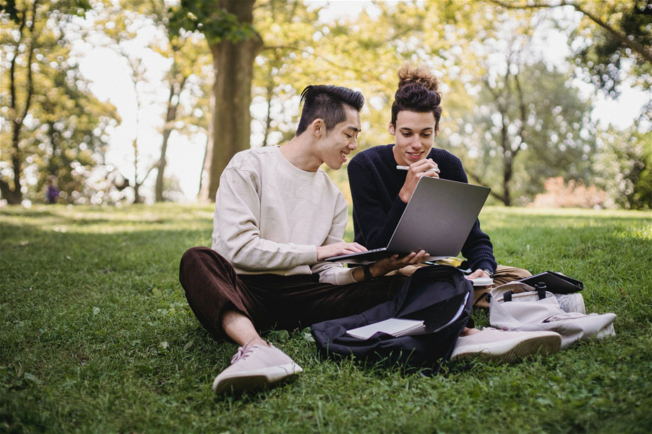 Two students sitting on grass happily working on computer