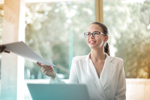 Smiling business woman at her desk in an office being handed a small stack of documents