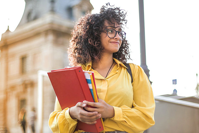 female student in a yellow spring jacket and glasses holding books looing on optimistically