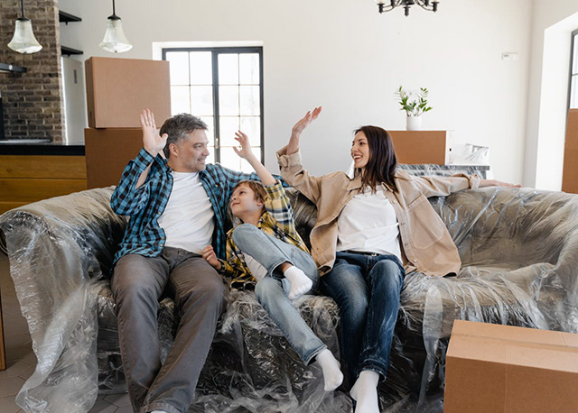 Father mother and son sitting and high fiving each other  on a wrapped couch in a new home with moving boxes in the room