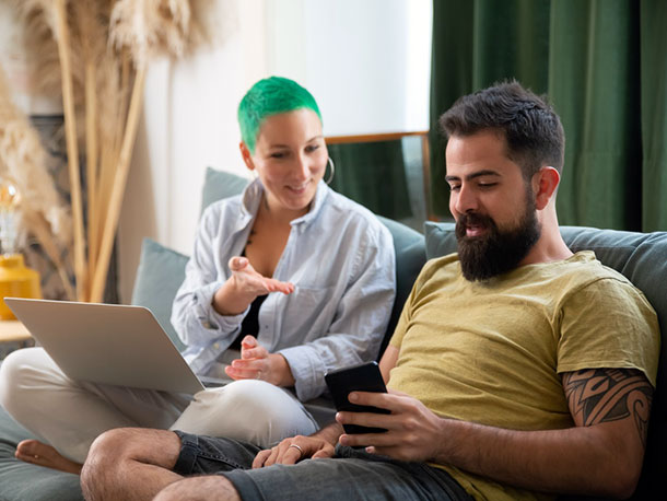 Man using a phone Sitting Beside woman using a laptop on a couch at home Man using a phone Sitting Beside woman using a laptop on a couch at home
