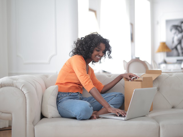 Cheerful woman sitting on couch opening box and using laptop Cheerful woman sitting on couch opening box and using laptop
