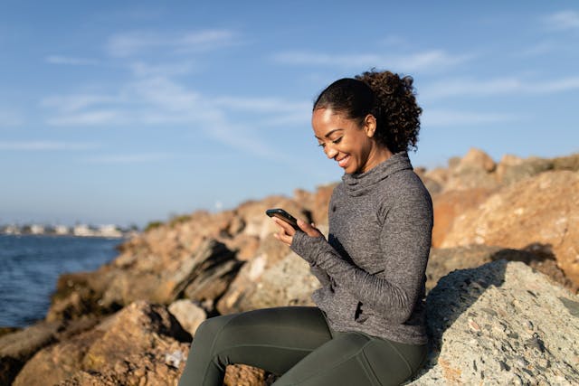 Female sitting on a rocky beach happily using her smartphone