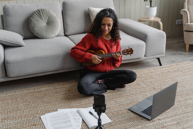 Female sitting on the floor in a living with a laptop on sheet music practicing her ukulele